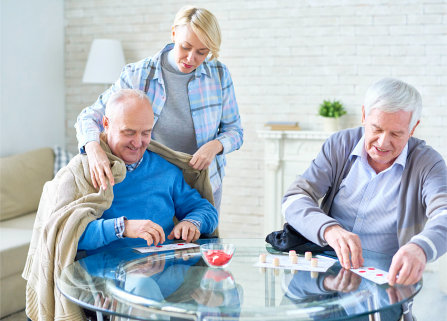 two seniors men busy playing cards while caregiver is putting a blanket to a senior man